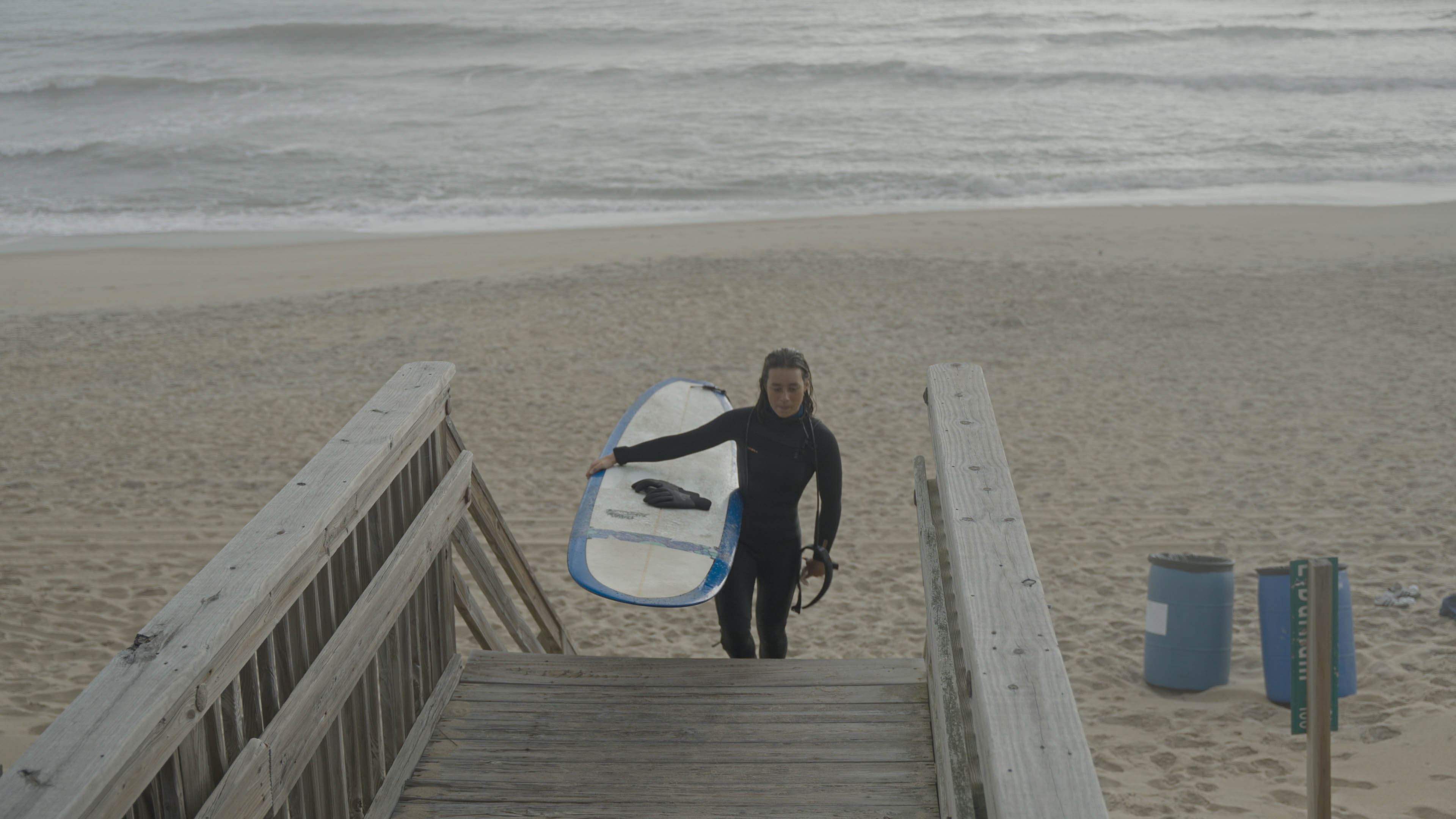 A young woman walks up a set of stairs holding a surfboard as beach waves crash behind her.