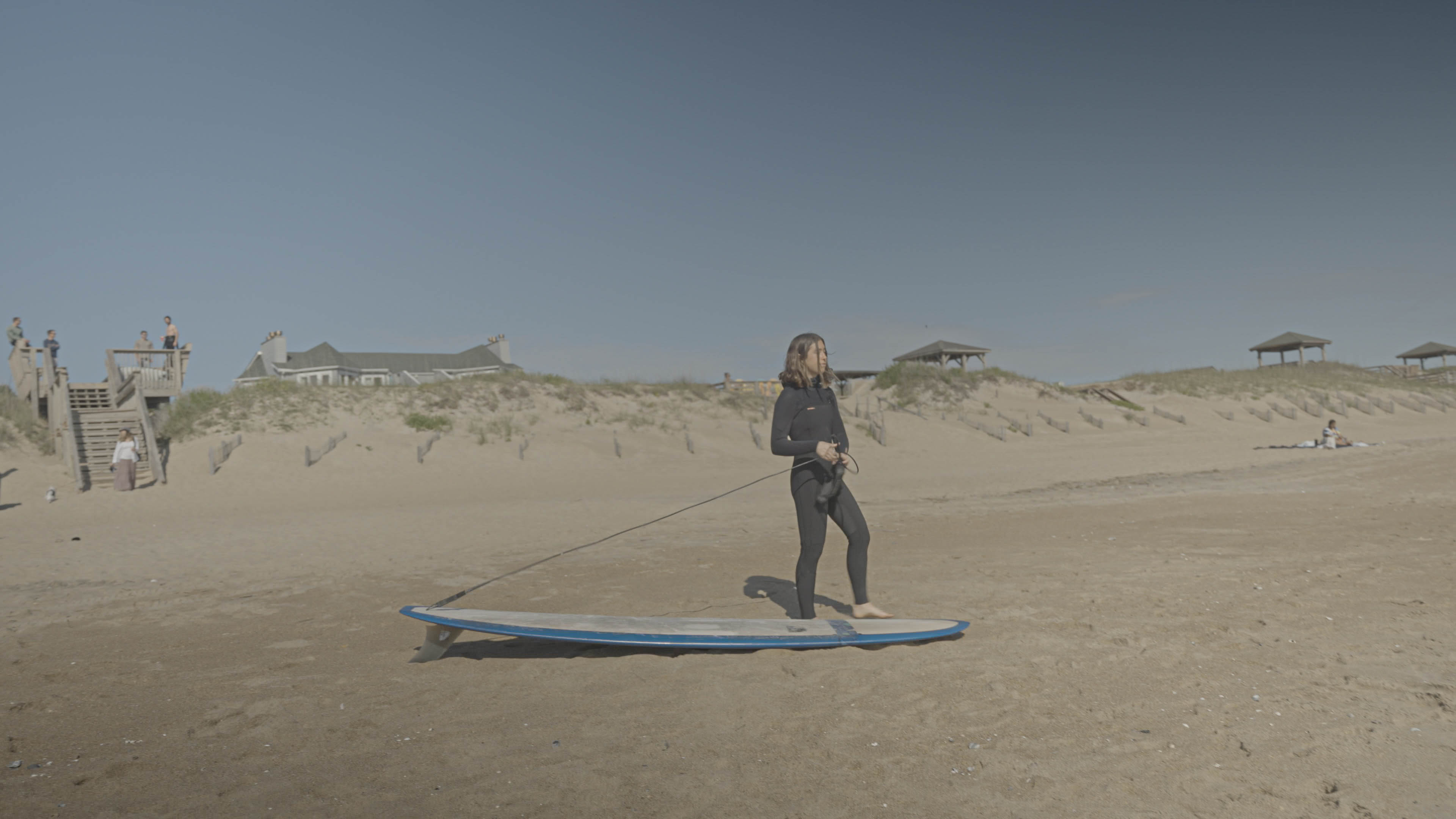 A young woman looks off into the distance in front of a blue surfboard.