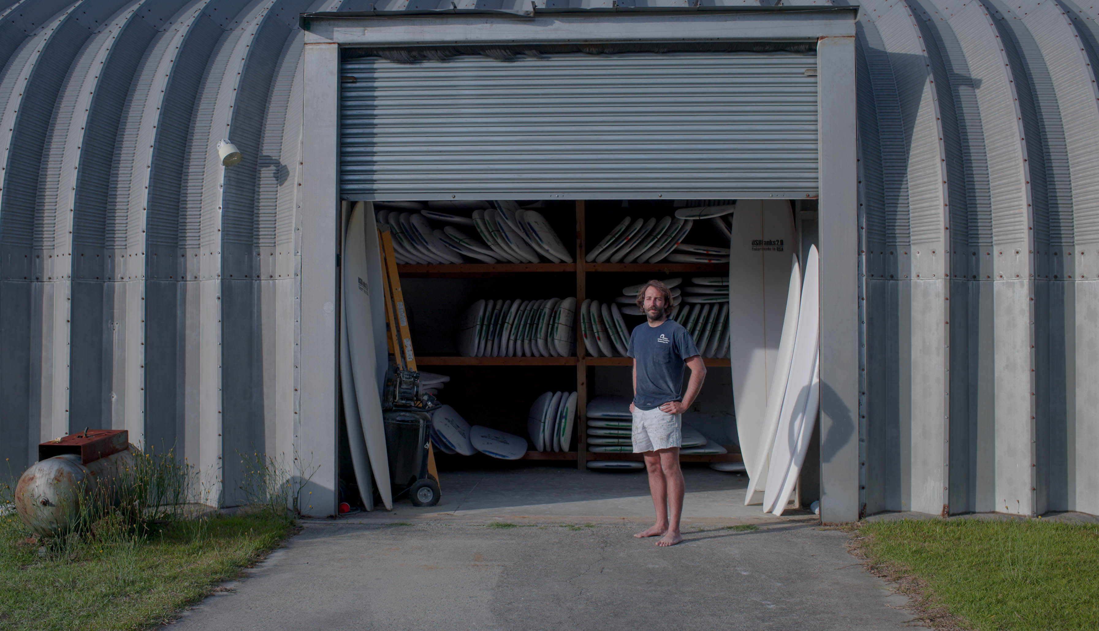 A young man stands in front of garage door, blank surfboards sit behind him.