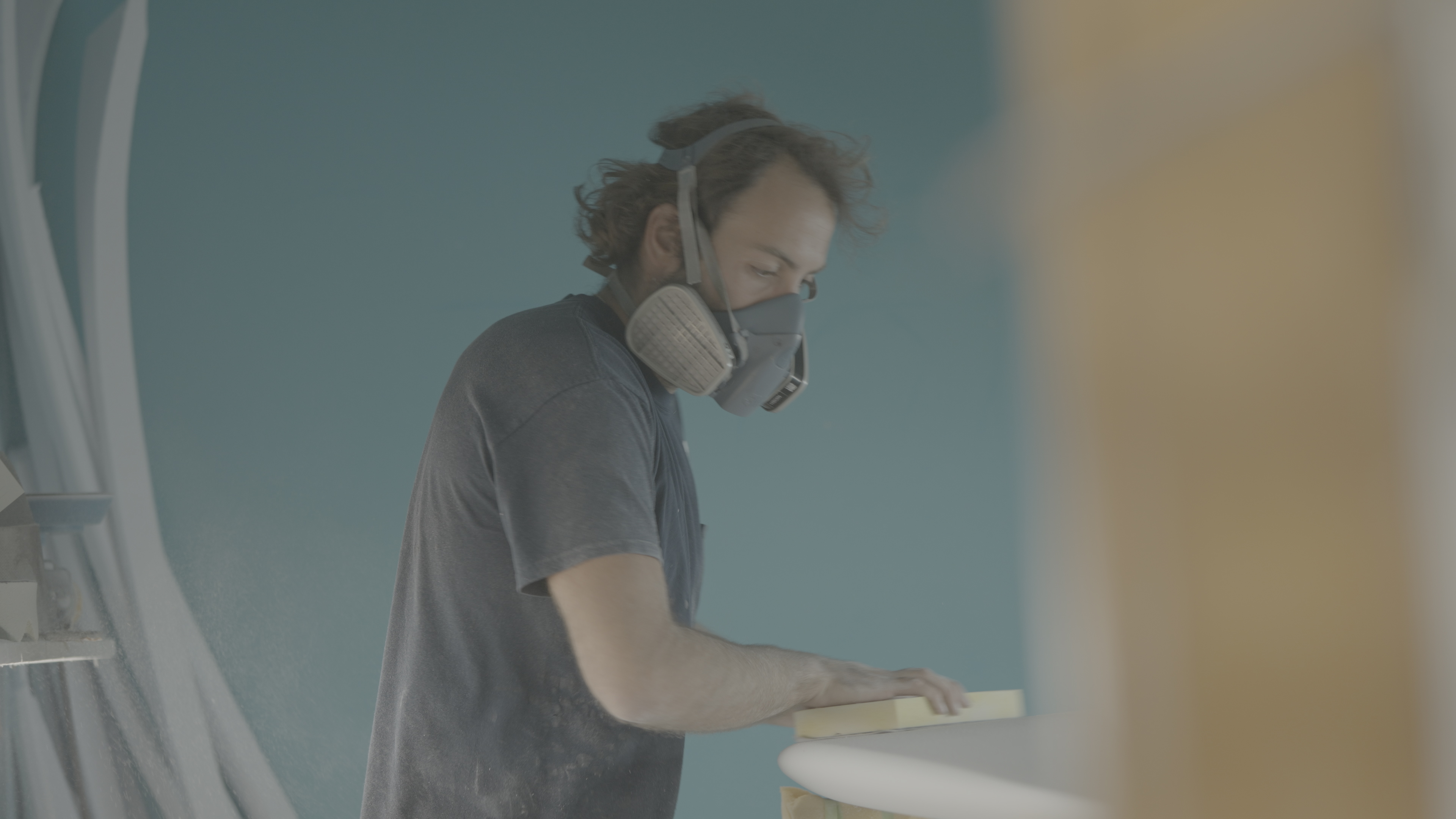 A young man wearing a construction masks, uses a shaving tool to shape a surfboard.