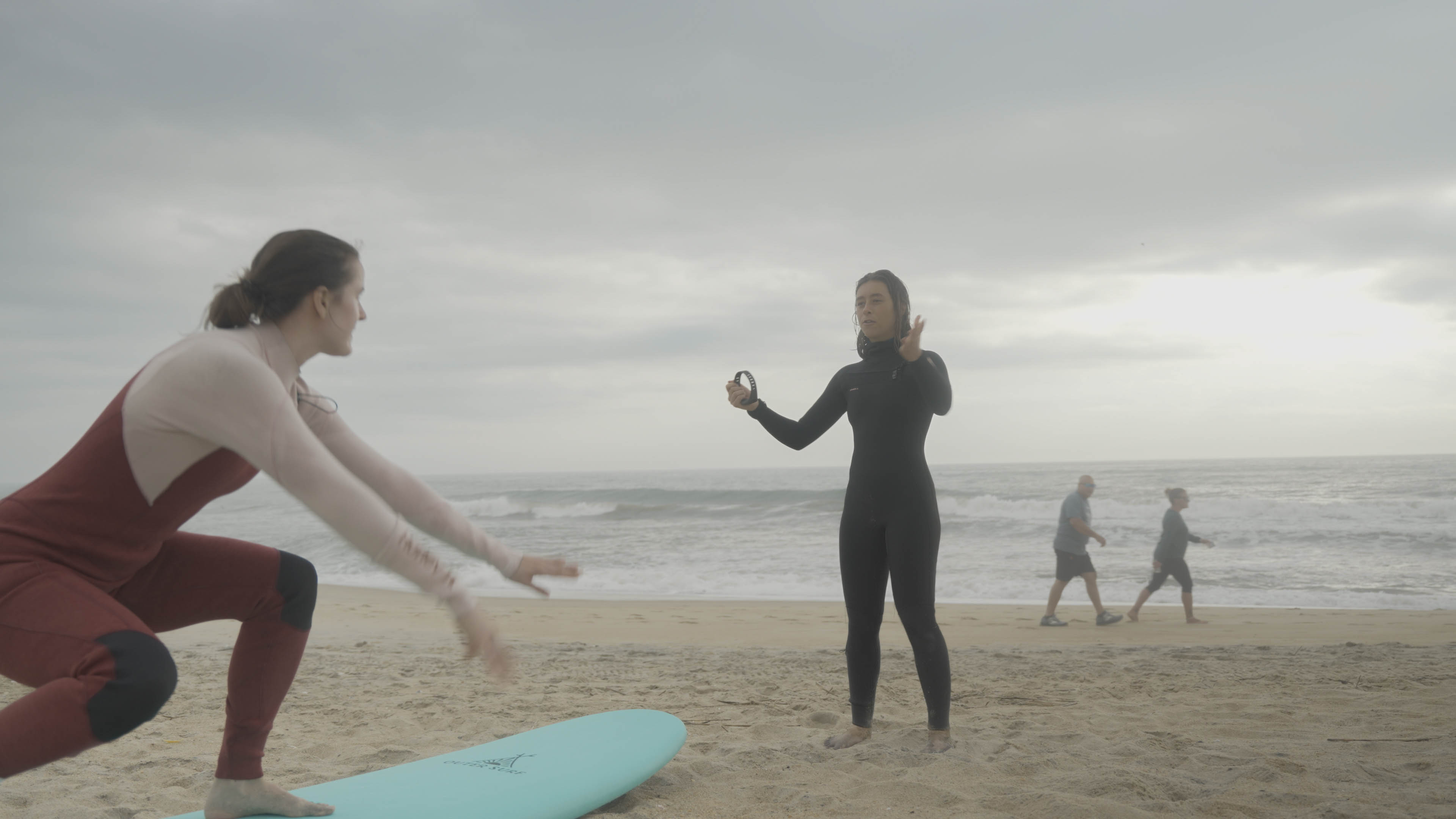 A young woman in surf gear stands in front of an older woman as she practices balancing on a surfboard.
