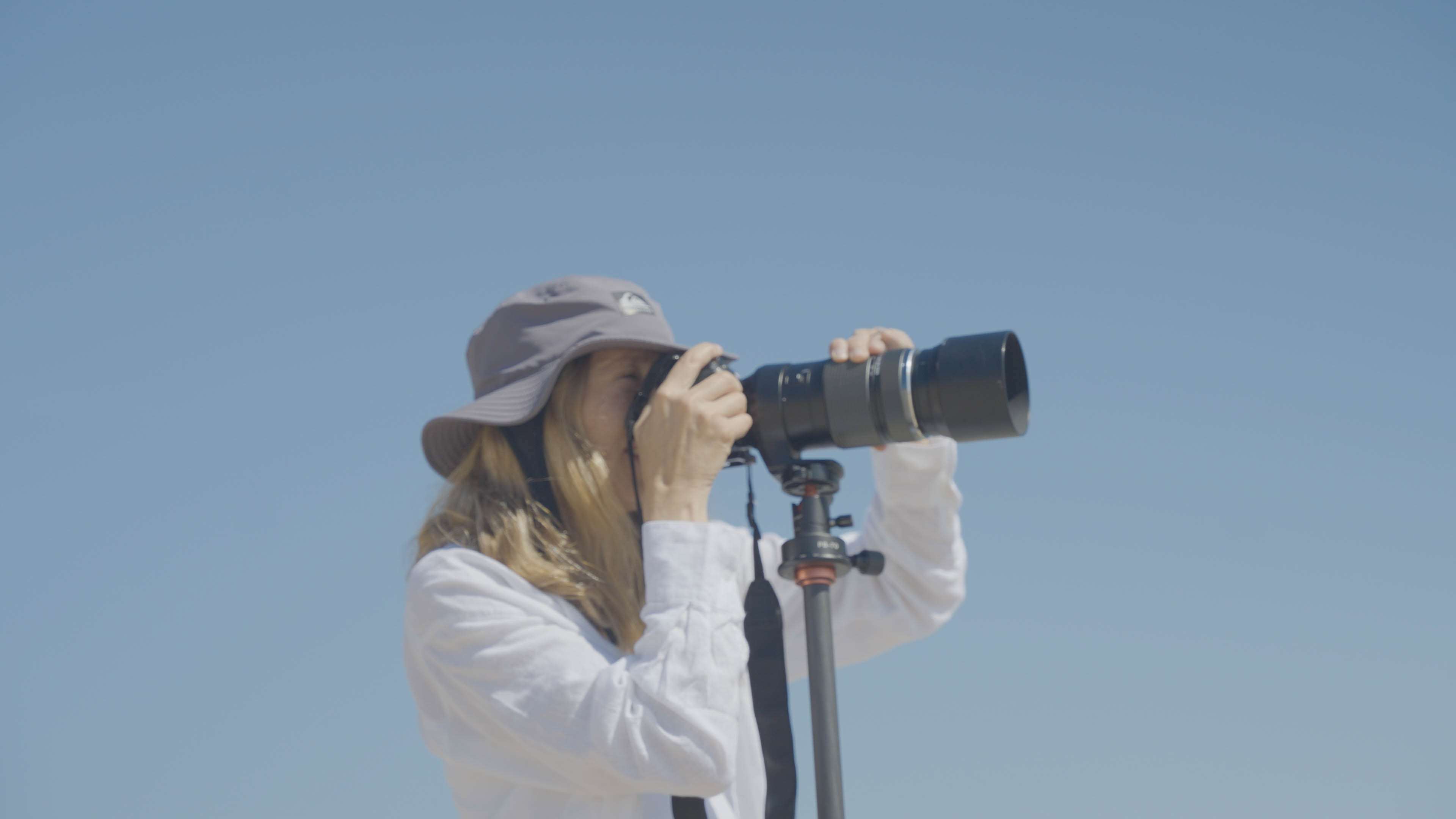 An older woman is looking in the viewfinder of a digital camera, with only blue skies behind her.