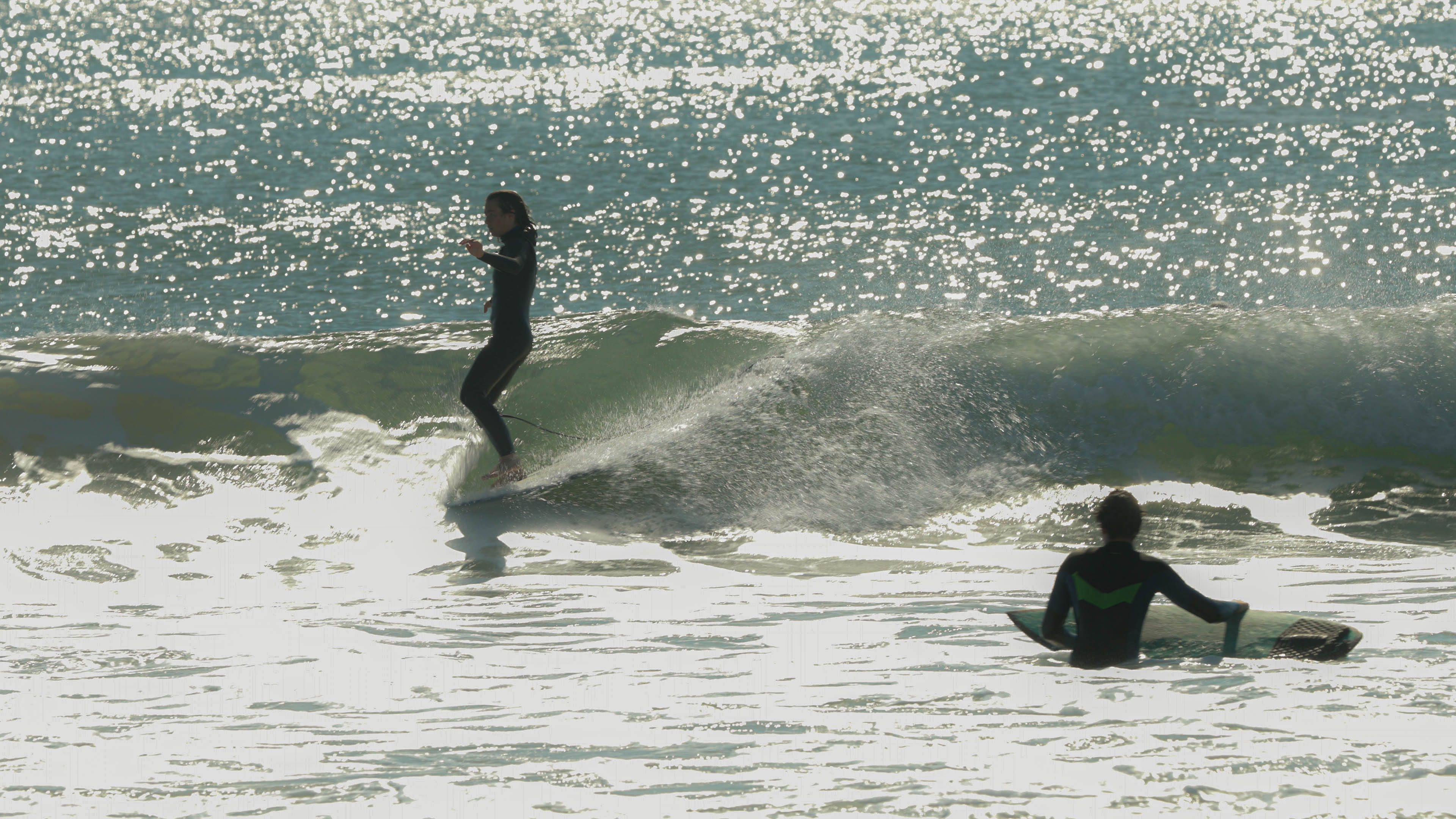 A young woman rides a wave on a surfboard while another person in the water watches.
